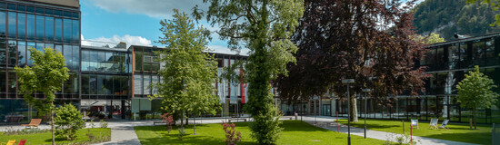 Campusgarten mit Sitzmöglichkeiten vor dem modernen Gebäude der FH Kufstein Tirol Grüne Außenanlage mit Bäumen und Wegen vor einem modernen Hochschulgebäude an einem sonnigen Tag