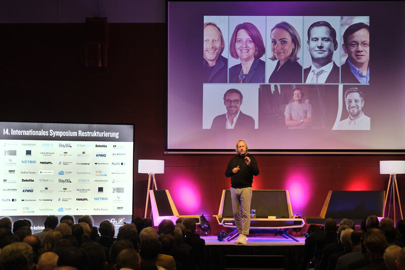 Markus Exler, Gastgeber des 14. Symposiums Restrukturierung auf der Bühne. Im Hintergrund ein Screen mit den Porträts der Teilnehmenden der Paneldiskussion, Stehlampen , eine Sitzgruppe und eine Sponsorenwand mit Logos