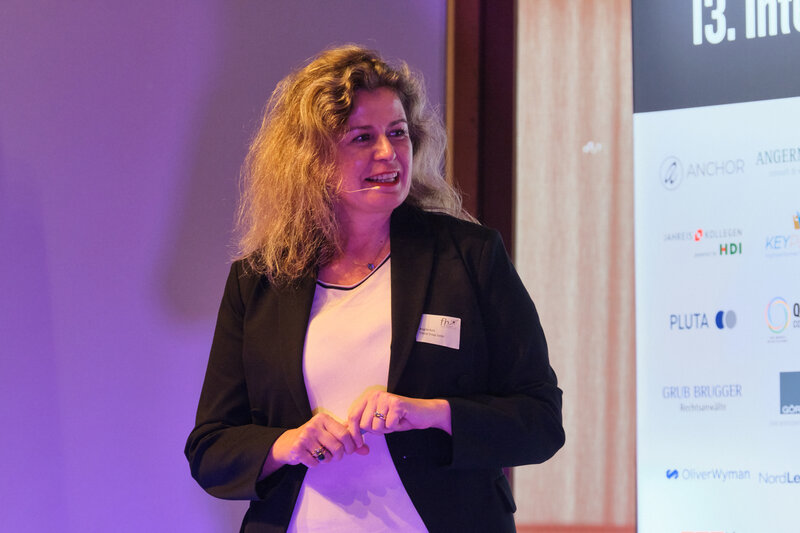 2024_Kurz | © FH Kufstein Tirol Eine Frau steht auf einer Bühne und hält einen Vortrag, im Hintergrund sieht man Sonsorenlogos und den Schriftzug "13. Internationales Symposium Restrukturierung" | © FH Kufstein Tirol