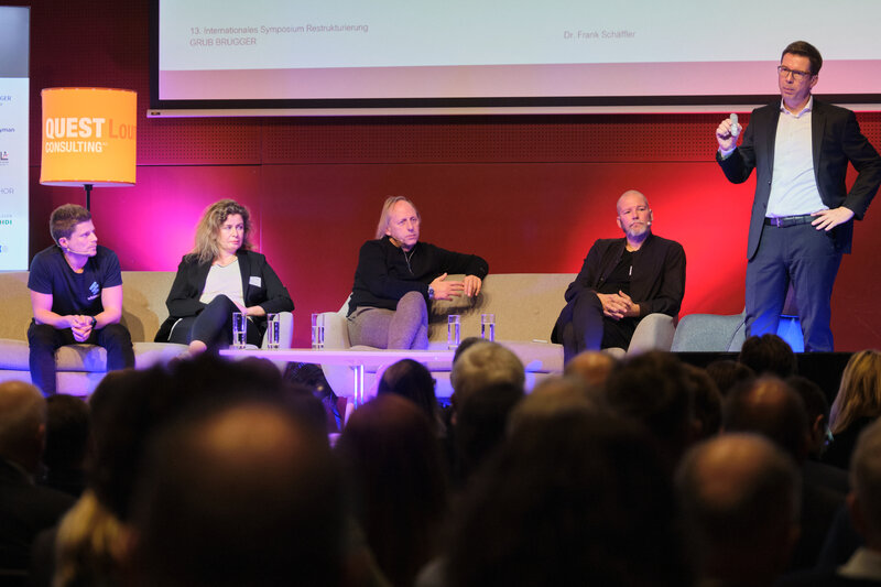 2024_Schaeffler | © FH Kufstein Tirol Diskussionsrunde auf einer Bühne, ein Mann mit Headset hält einen Vortrag, im Vordergrund sind die Kopf von Teilnehmer:innen aus dem Publikum zu sehen, auf der Bühne verfolgen eine Frau und drei Männer den Votrag | © FH Kufstein Tirol