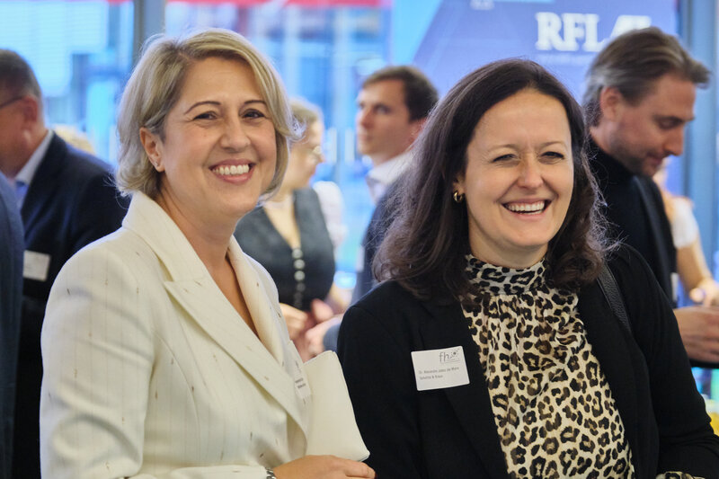 2024_Incoronata_Alexandra Zwei Frauen in Businesskleidung auf einer Veranstaltung lächeln in die Kamera, weitere Personen stehen im Hintergrund