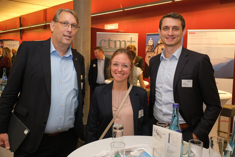 2024_VTR | © FH Kufstein Tirol ein Mann und eine Frau stehen bei einer Veranstaltung an einem Stehtisch, im Hintergrund sind weitere Personen zu sehen, außerdem Aufsteller mit Firmennamen und Logos | © FH Kufstein Tirol