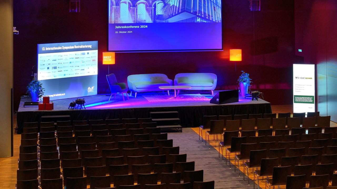 2024_Buehne | © FH Kufstein Tirol Leerer Konferenzsaal mit Bühne, moderner Möblierung und Präsentationsbildschirm, vorbereitet für die Jahreskonferenz 2024. | © FH Kufstein Tirol
