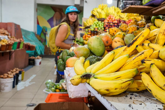 Im Vordergrund Berge von Bananen, Mangos und Trauben - Eva beim Einkaufen im Hintergrund