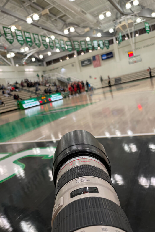 Objektiv einer Kamera im Hintergrund ein Basketball-Spielfeld