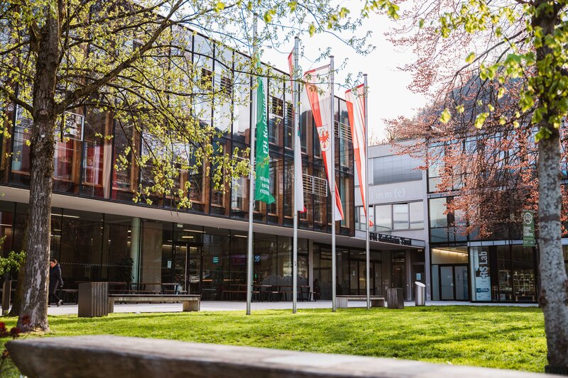 Frühlingsstimmung vor dem Haupteingang der FH Kufstein Tirol Außenansicht der FH Kufstein Tirol mit Fahnen vor einer modernen Glasfassade und Frühlingsbäumen