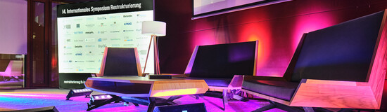 Bühne der 14. Jahreskonferenz Restrukturierung an der FH Kufstein Tirol Bühne der 14. Jahreskonferenz Restrukturierung an der FH Kufstein Tirol, eine moderne Sitzgruppe und mit Tisch und Stehlampe, dahinter eine Aufsteller mit dem Schriftzug 14. Internationales Symposium Restrukturierung und vielen Logos. Die Bühne wird von lila- rosa-farbigen Scheinwerfern angestrahlt